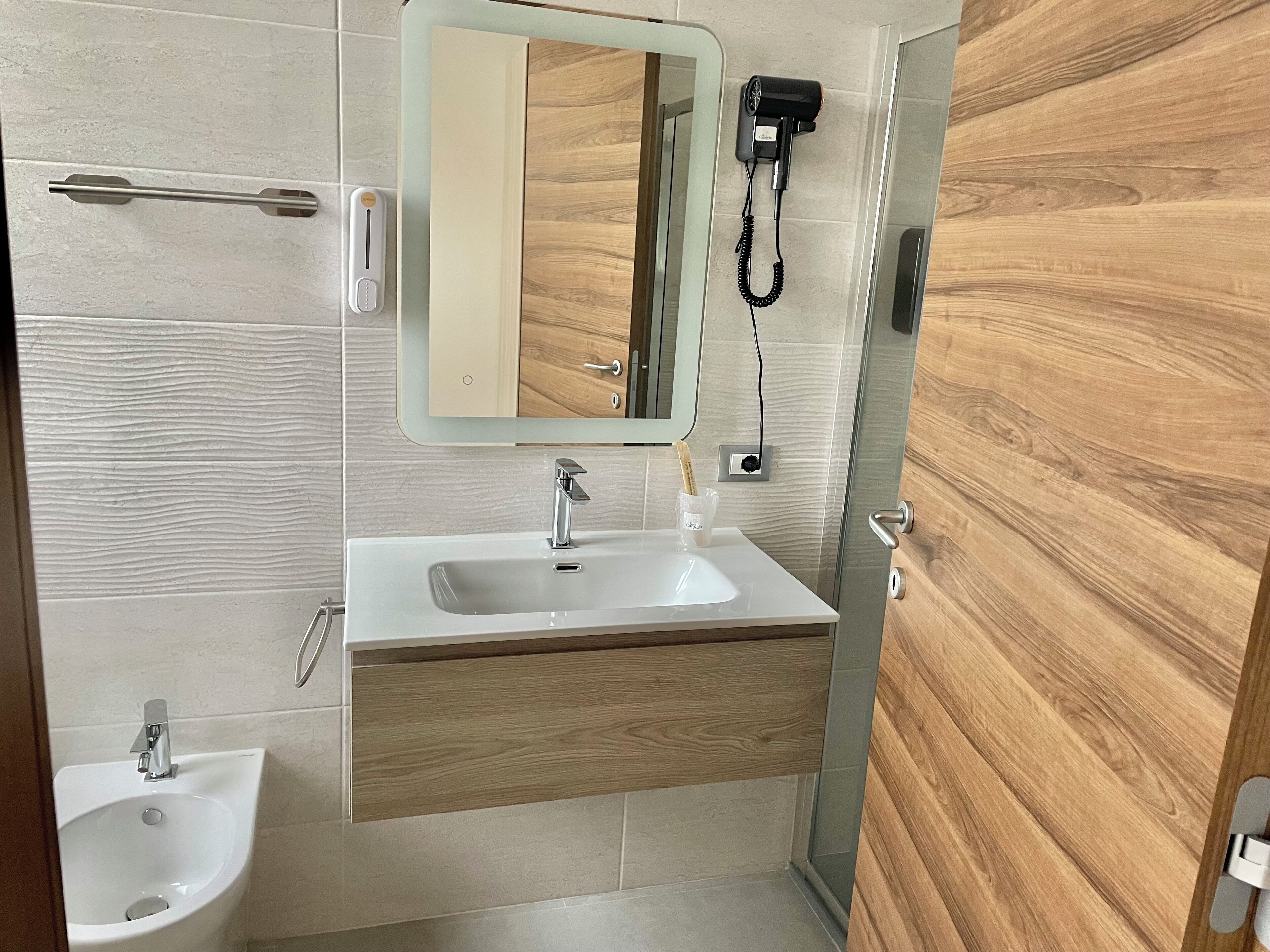 A modern bathroom with a white sink, a bidet, a wooden vanity, a mirror, and a hairdryer mounted on the wall.