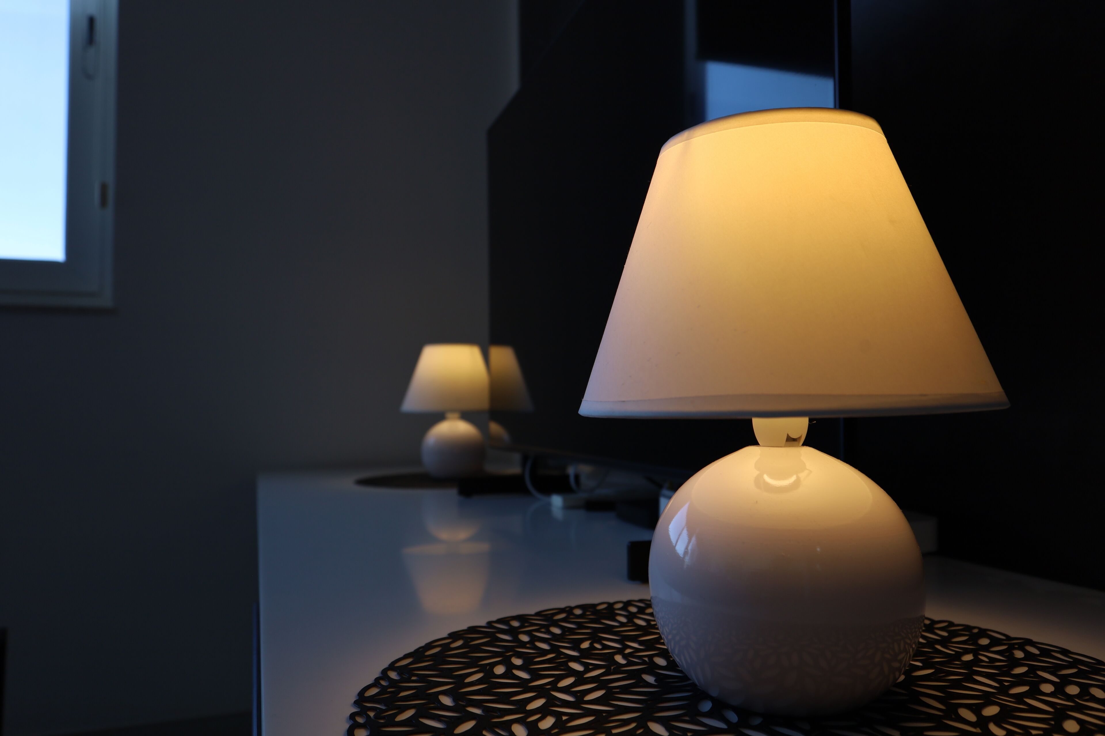 A modern lamp on a table with a patterned coaster, a television, and a wall-mounted lamp in a dimly lit room.