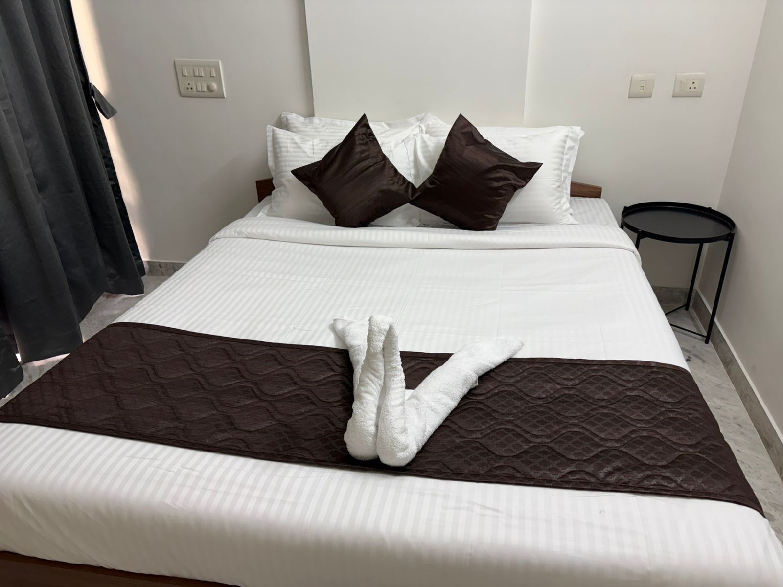 Una cama bien hecha con ropa de cama blanca y marrón, una mesita redonda a un lado y una cortina oscura parcialmente visible.
