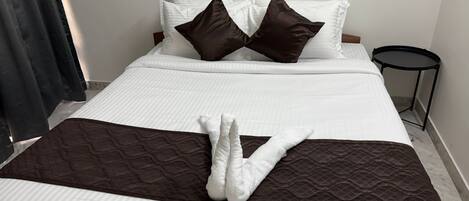 Una cama bien hecha con ropa de cama blanca y marrón, una mesita redonda a un lado y una cortina oscura parcialmente visible.