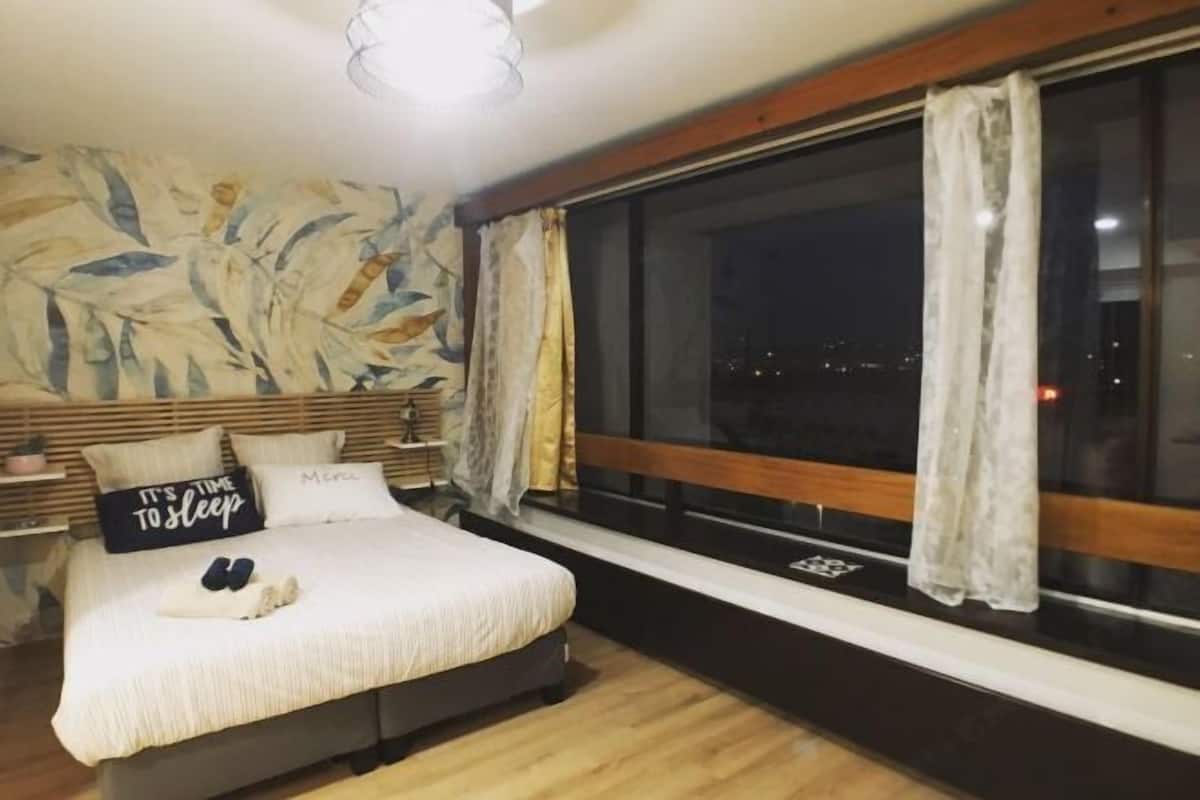 Ein Schlafzimmer mit Bett, Kissen, einem Fenster mit Vorhängen, einem Nachttisch und einer Wand mit Blätermotiv.