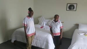Two individuals in a hotel room with two beds, a framed picture on the wall, and a bedside table with a lamp. - Zaru Hotel (Gizo)