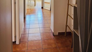 Un corridoio con pavimento piastrellato, una porta bianca, una scala appoggiata al muro e un frigorifero sullo sfondo.