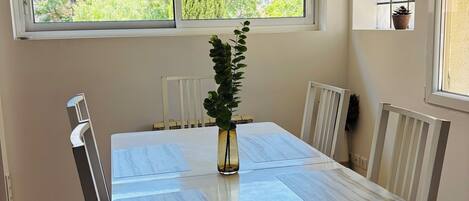 Un coin repas avec une table blanche, des chaises, un vase avec une plante et des fenêtres donnant sur un paysage verdoyant.