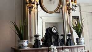 A room with a fireplace, a mantel with decorative items, and a mirror with a framed portrait. - Suite BORDEAUX park view (Hastingues)