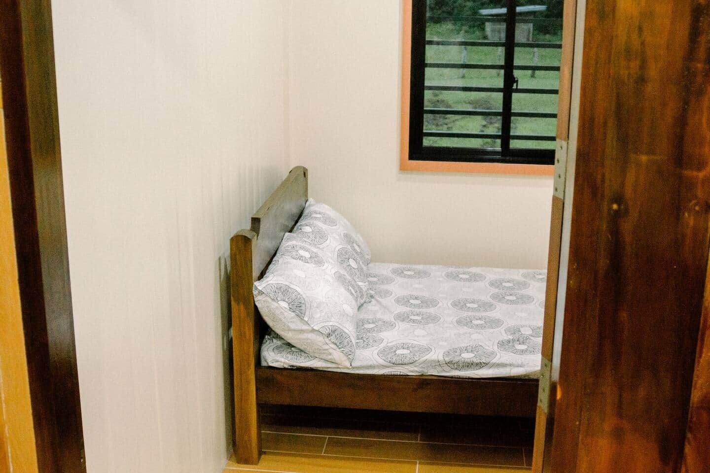 Una habitación pequeña con cama individual, cabecera de madera, colchón estampado y ventana con persianas horizontales.