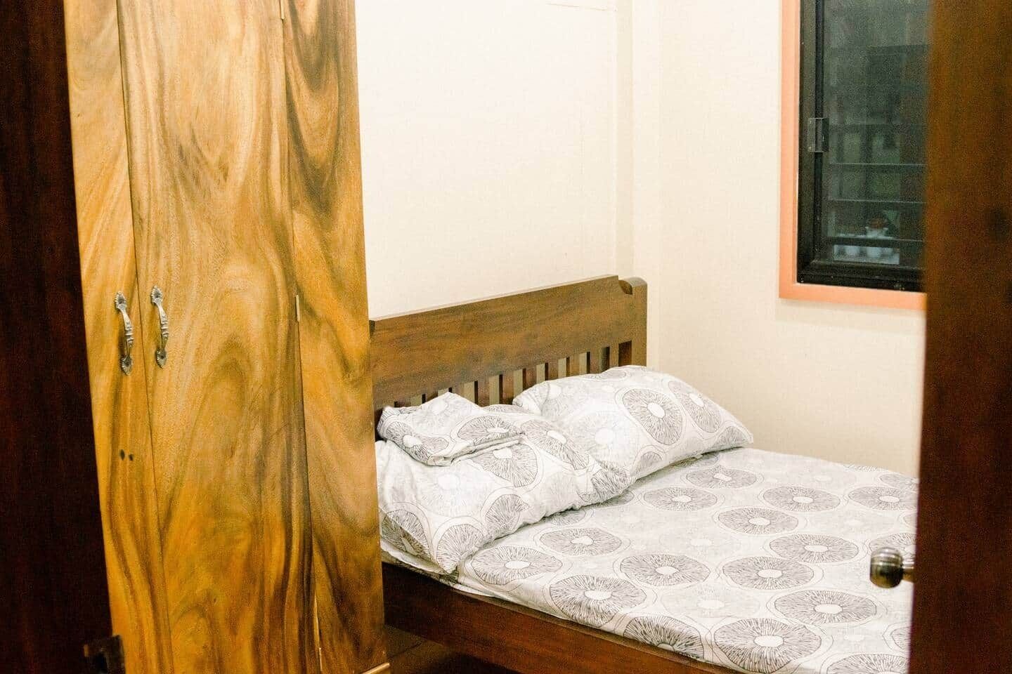 A small bedroom with a wooden wardrobe, a single bed with patterned bedding, and a window with a view of buildings.