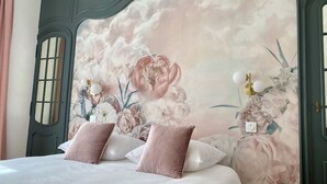 Une chambre avec un grand décor mural floral sur le mur, un lit avec une literie blanche et rose, et une porte de style ancien dotée d’une fenêtre.