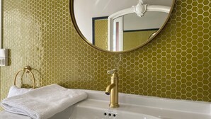 Une salle de bain avec un mur recouvert de carreaux hexagonaux dorés, un lavabo blanc, un robinet doré et un miroir rond.