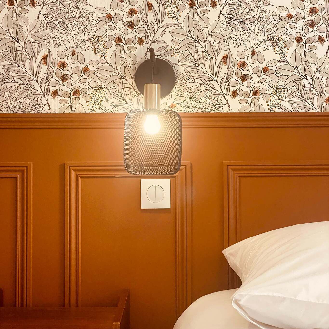 Ein Zimmer mit floralem Wandpapier, einer Wandleuchte und einem Bett mit weißem Kopfkissen.