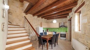 Una scala conduce a una sala da pranzo con tavolo e sedie in legno, una cucina sullo sfondo e pareti in pietra.