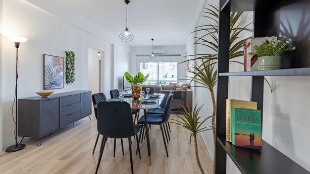A modern dining area with a wooden floor, black chairs, a central table with a vase and glassware, and a bookshelf with books and plants. - Citrus 3-BR Apt. & Balcony in Larnaca (Larnaca)