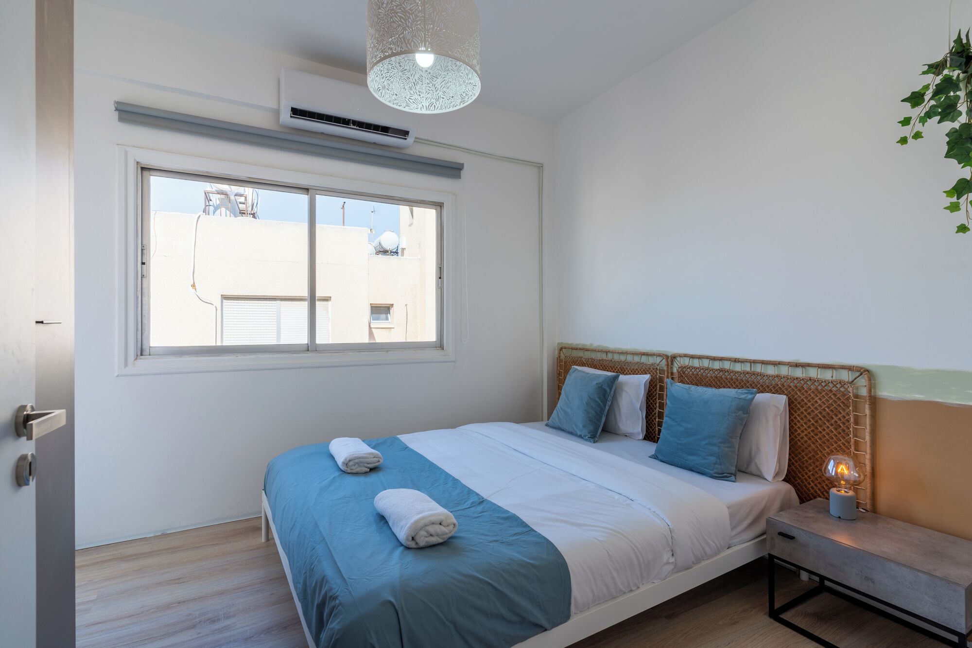 Un dormitorio con cama, ventana, equipo de aire acondicionado y mesita de noche con lámpara.