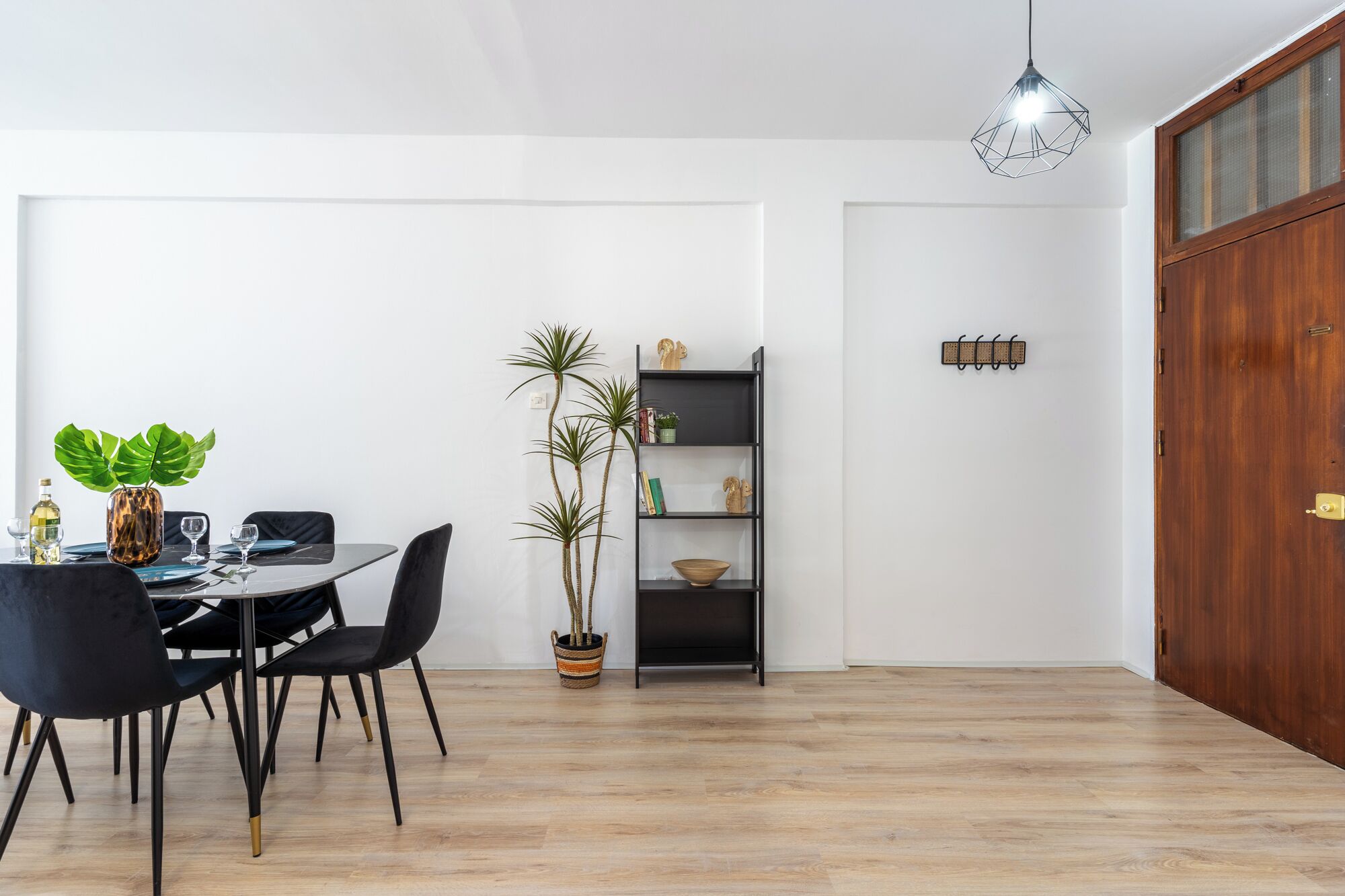 A dining area with a wooden table, black chairs, a potted plant, and a black shelving unit against a white wall.