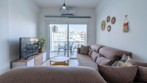 A modern living room with a sofa, a TV, a small table, and a balcony with chairs. - Citrus 3-BR Apt. & Balcony in Larnaca (Larnaca)