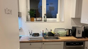 A compact kitchen with white cabinets, a dishwasher, a sink, and a window with a plant and a clock. - Beautiful, cozy accommodation with separate rooms, shared kitchen and bathroom (Langgöns)