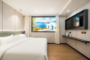 Une chambre d’hôtel avec un grand lit, un téléviseur fixé au mur, une fenêtre donnant sur la mer et une petite étagère avec quelques objets.