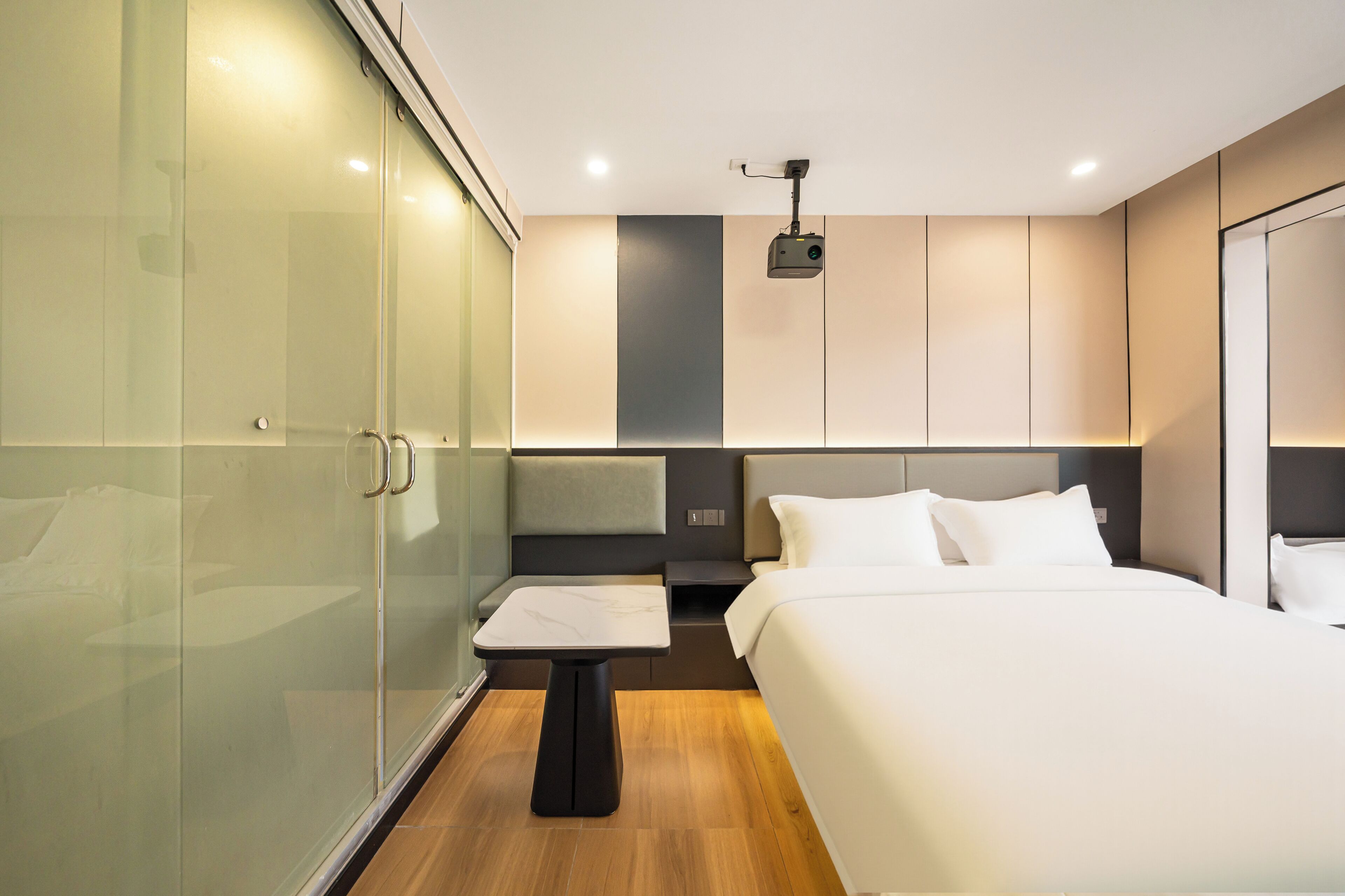 Ein modernes Hotelzimmer mit einer Glastruhe, einem Bett mit weißer Bettwäsche, einem kleinen Tisch und einem Deckenprojektor.