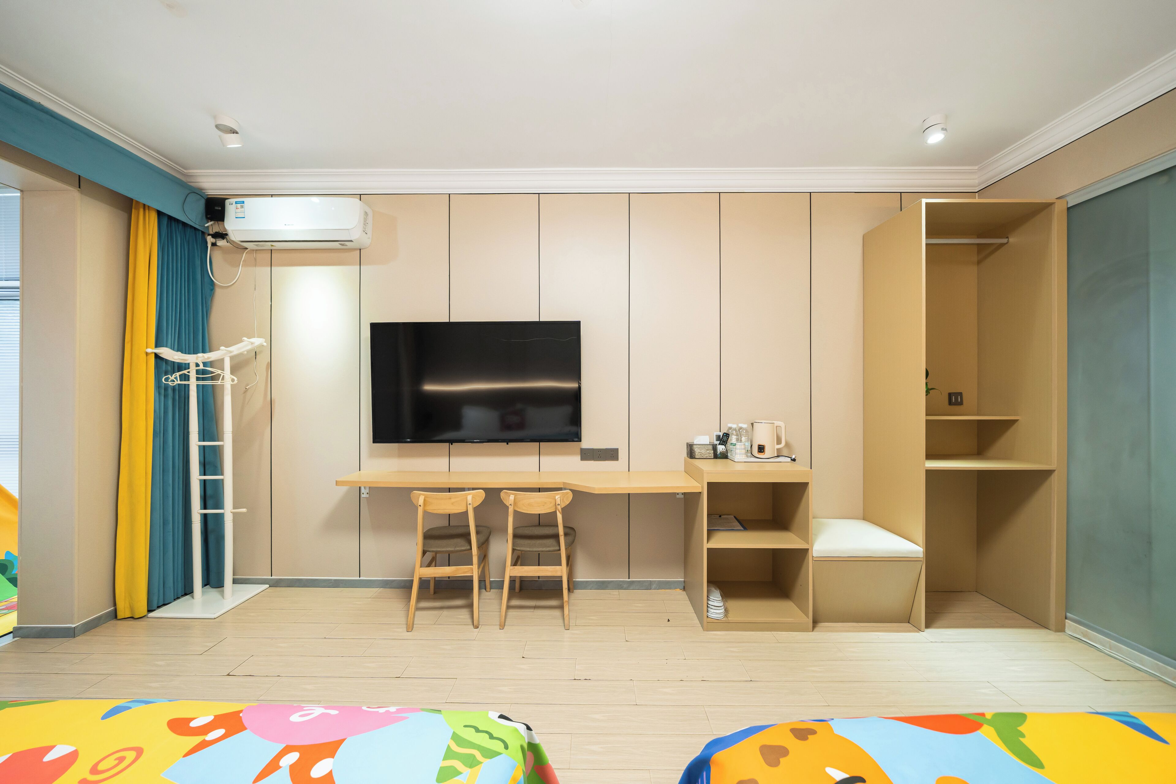 A modern room with a flat-screen TV mounted on the wall, a wooden desk with two chairs, a small seating area, and a wardrobe with a mirror.