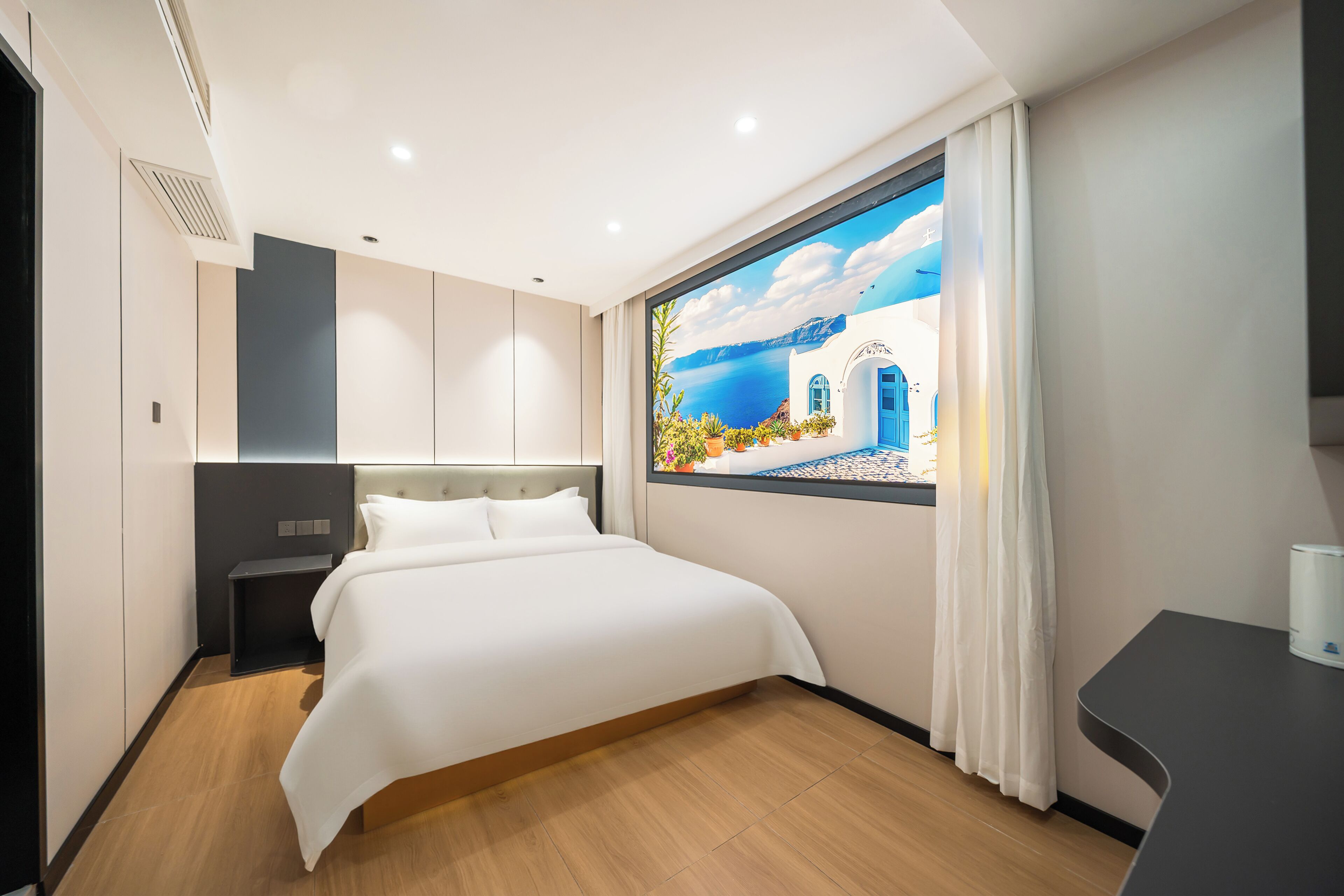 A modern hotel room with a large bed, a nightstand, a desk, and a view of a coastal scene through a window.