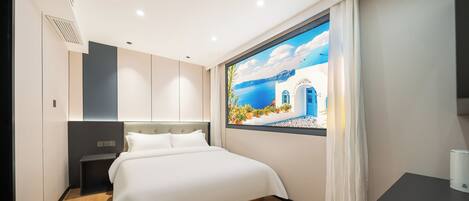 A modern hotel room with a large bed, a nightstand, a desk, and a view of a coastal scene through a window.