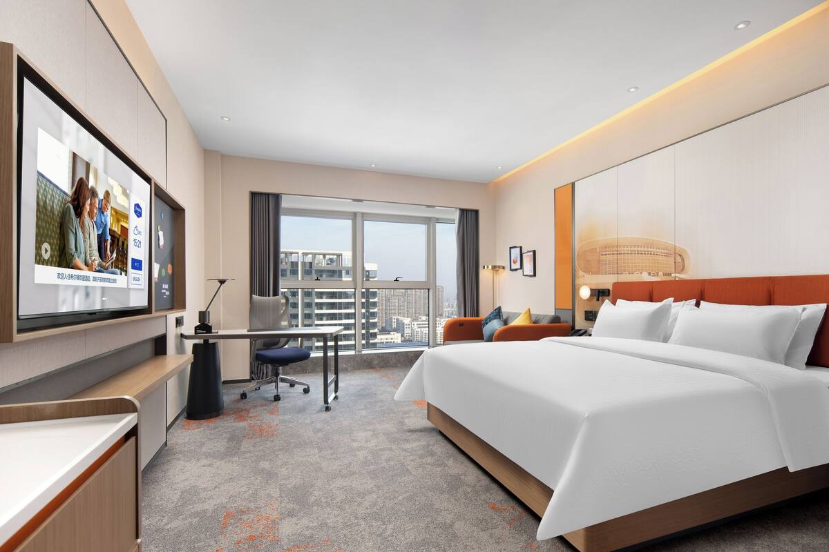 Habitación de hotel moderna con una cama grande, un escritorio con una silla, un televisor de pantalla plana y vistas a la ciudad a través de amplios ventanales.