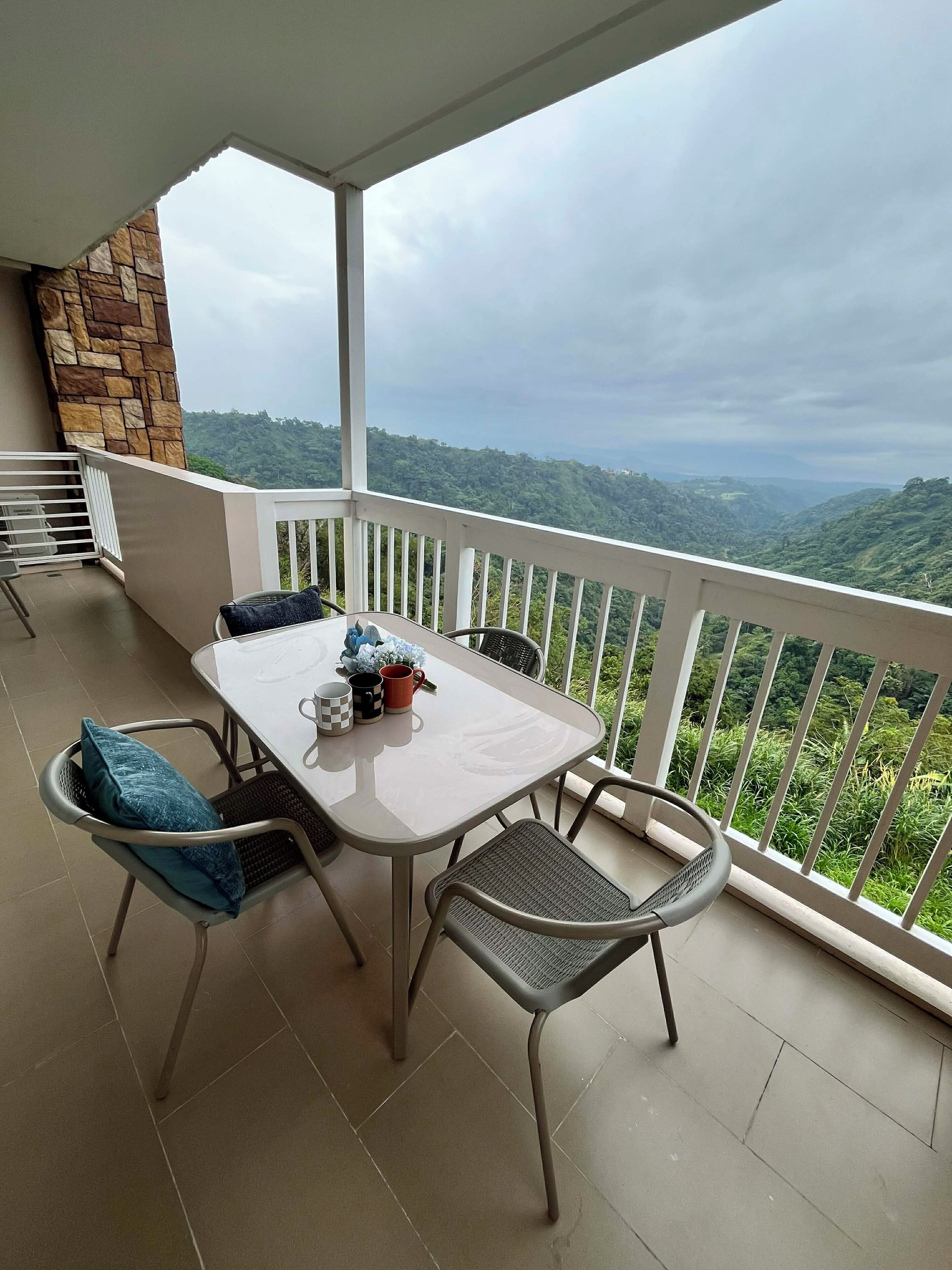 A balcony with a table and chairs, a stone wall, and a view of a green landscape.