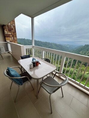A balcony with a table and chairs, a stone wall, and a view of a green landscape. - The Vineyard at Merlot TwinLakesTagaytay (Laurel)