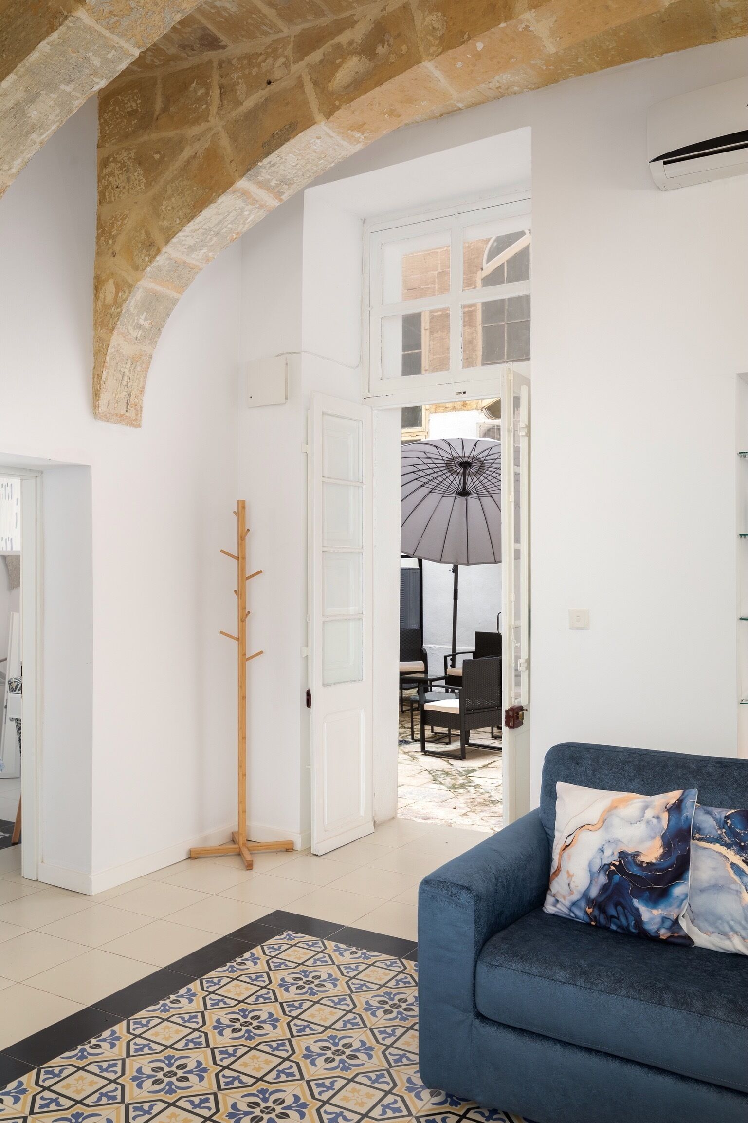 Ein Wohnzimmer mit blauem Sofa, einem gemusterten Teppich, einem Garderobenständer und Blick auf einen Außenbereich mit Essplatz durch eine offene Tür.