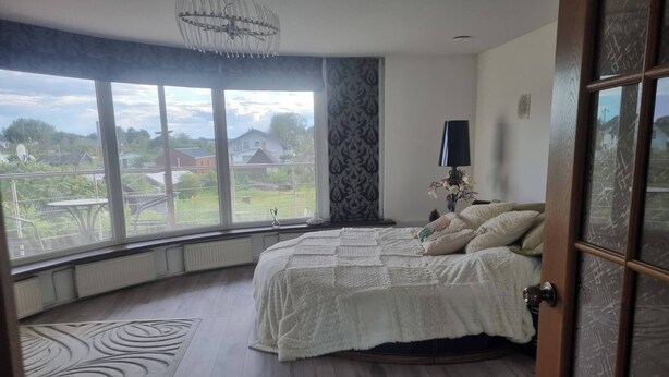 A bedroom with a large window, a bed with white bedding, a patterned rug, and a wooden floor.