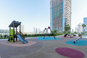 Ein Spielplatz mit Rutsche, Schaukeln und Wippe, umgeben von einer Grünanlage mit Palmen und Blick auf hohe Gebäude im Hintergrund.
