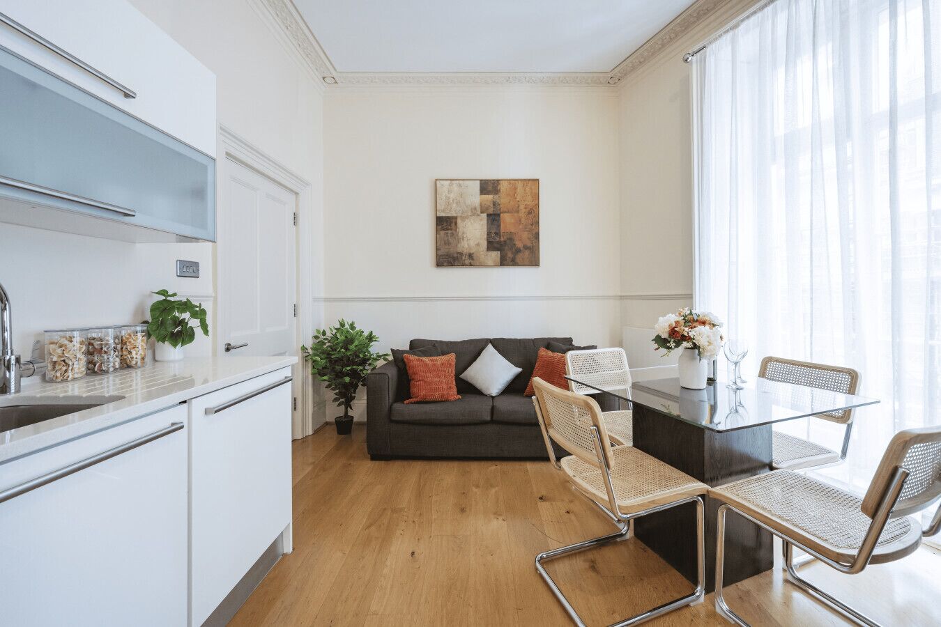 Una cocina moderna con armarios blancos, una zona de comedor con mesa de vidrio y sillas de mimbre, un sofá gris con cojines y un gran ventanal con cortinas translúcidas.