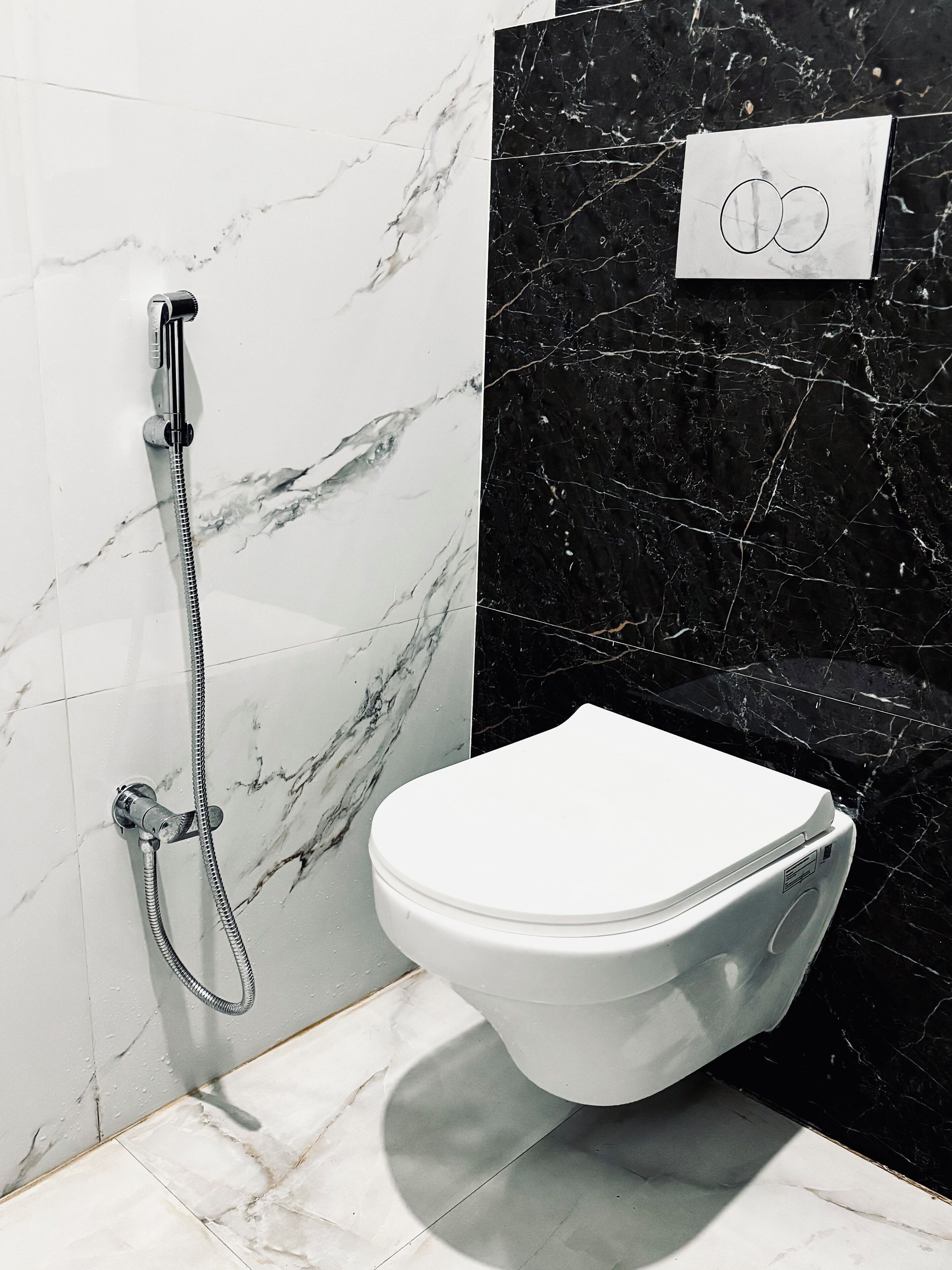 A bathroom with a white toilet, a shower with a silver hose, and marble-patterned walls.