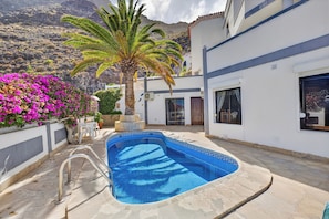 Ein Swimmingpool mit blau gefliestem Rand, eine Palme und rosafarbene Blumen in einer Wohngegend.