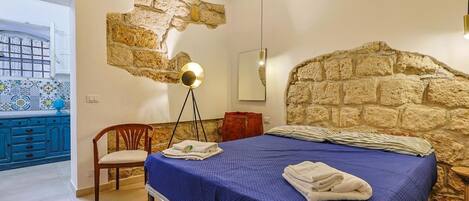 A bedroom with a stone wall, a bed with a blue bedspread, a chair, a bedside table, and a lamp.