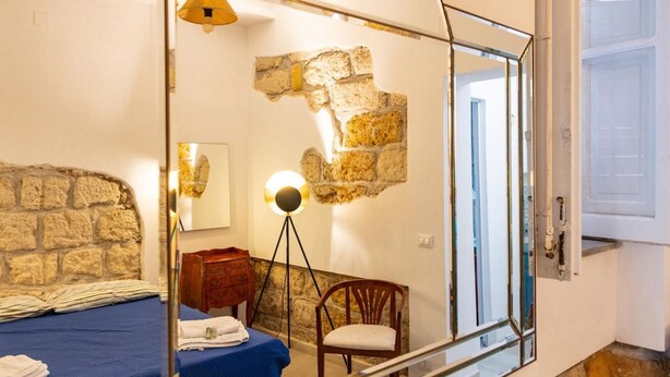 Un dormitorio con pared de piedra, una cama con colcha azul, una silla de madera, una lámpara de pie y un espejo grande.