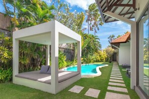 A modern outdoor seating area with a pool, surrounded by lush greenery and a clear sky. - Villa Alice Satu (Bali)