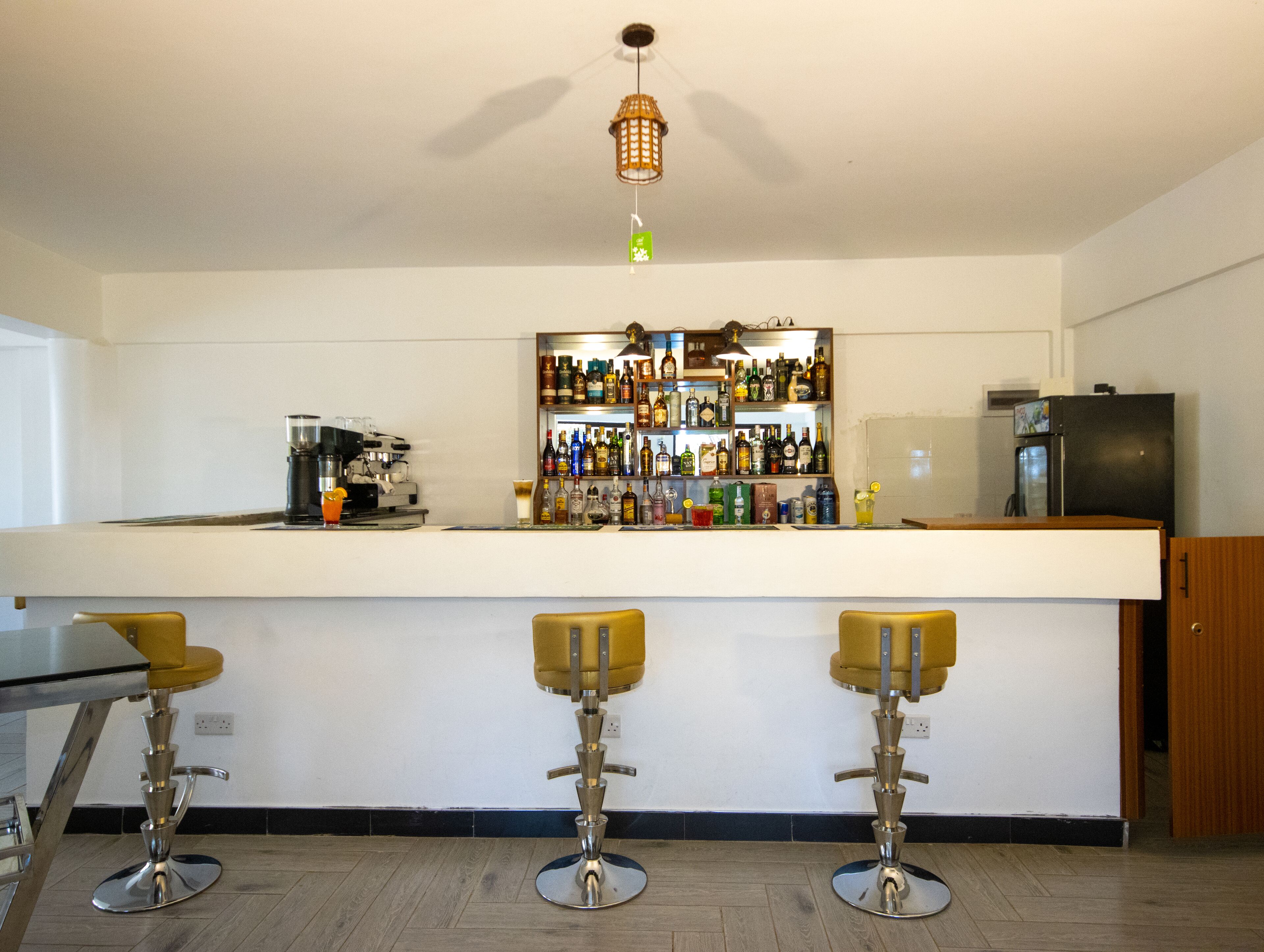A bar area with a counter, three stools, a coffee machine, and shelves with bottles.