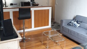 Ein modernes Wohnzimmer mit Holzboden, einem Glastisch, einem grauen Sofa mit einem gemusterten Kissen sowie einer Küchenzeile inklusive Mikrowelle und Spüle.