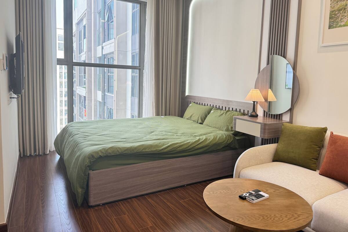 Une chambre d’hôtel avec un lit, un canapé, une table ronde en bois, une lampe, un miroir et une fenêtre avec des rideaux.