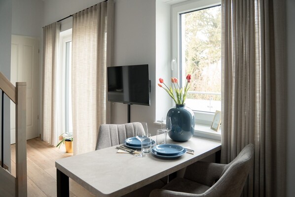 A dining area with a table set for two, a vase with flowers, a television, and a window with curtains.