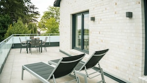 A balcony with two lounge chairs, a table, and chairs, overlooking a white brick building with a glass door. - Newly Built Wattblick Apartment in Dangast with Sea View, Large Private Roof Terrace & Loft (Varel)