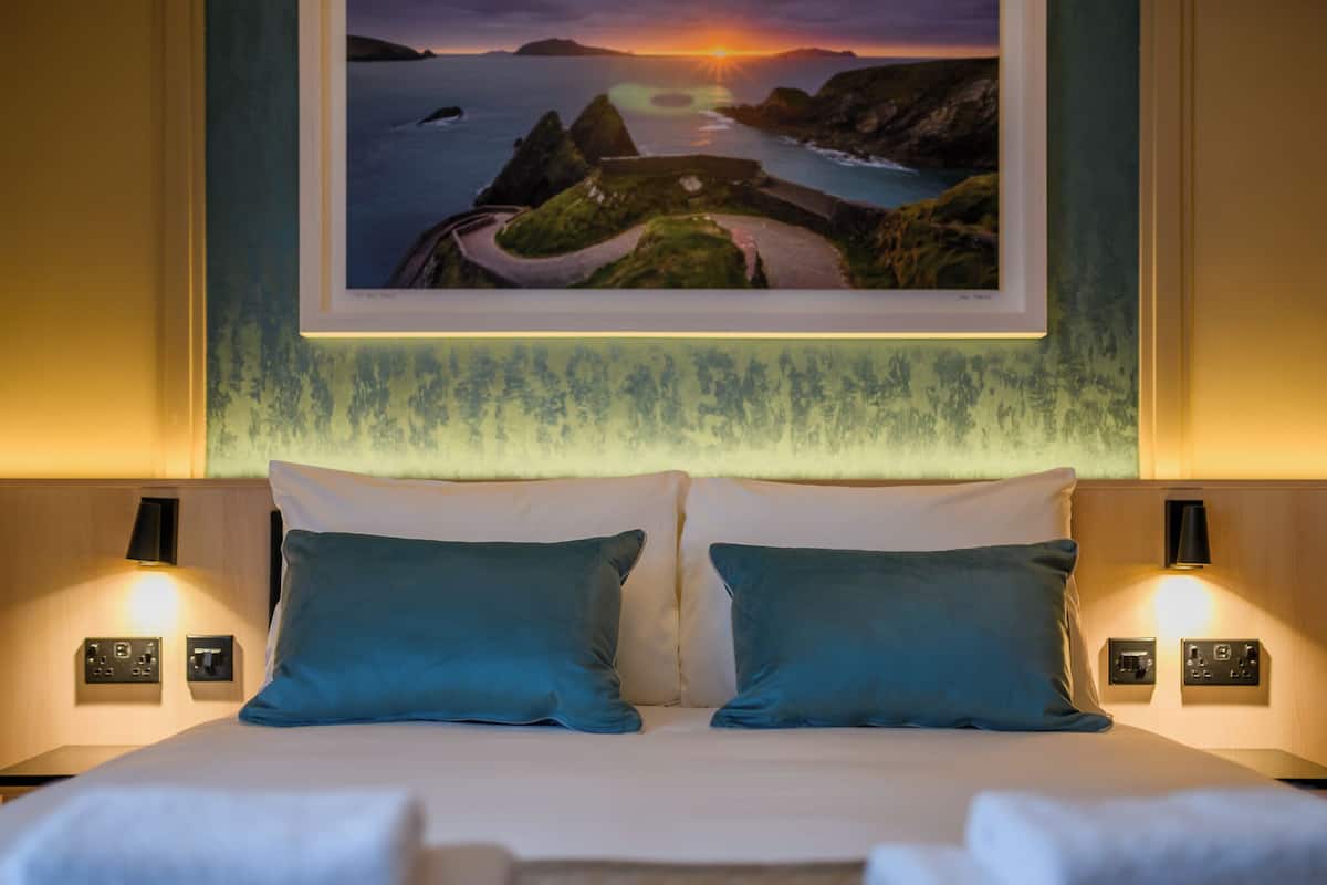 A hotel room with a bed, two pillows, bedside lamps, a wall-mounted picture, and a wall panel.