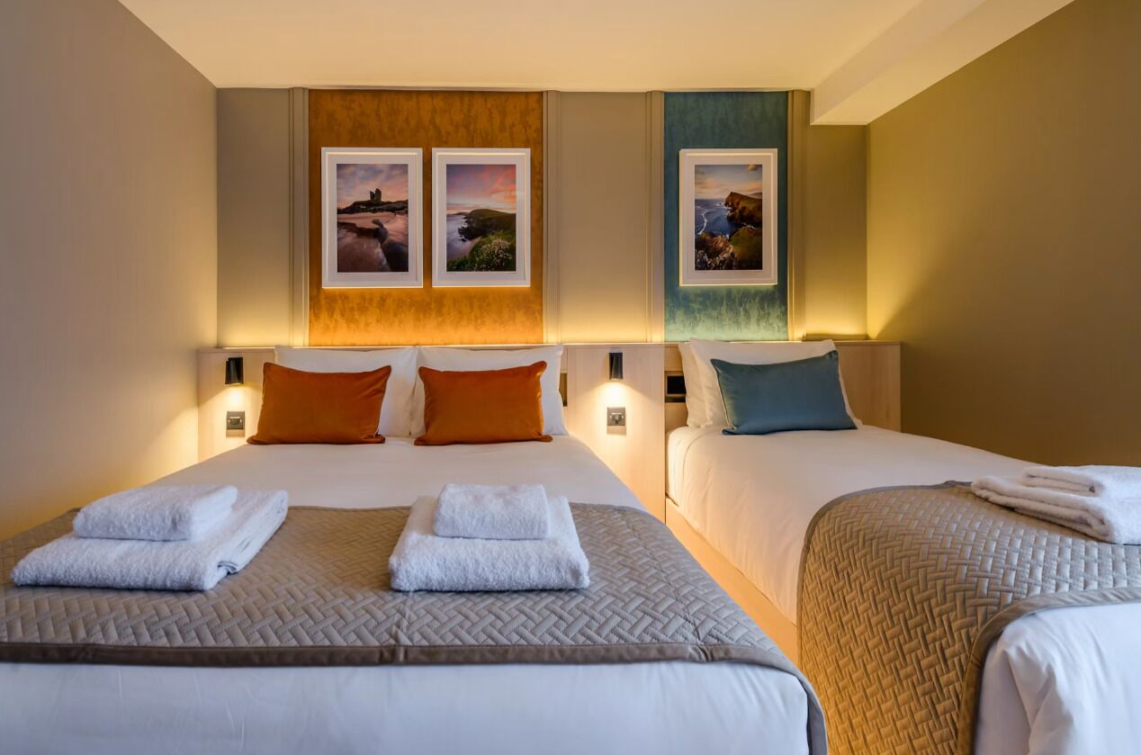 A hotel room with two beds, each with a headboard, white linens, and a quilted bedspread. There are two wall-mounted picture frames with landscape photos.