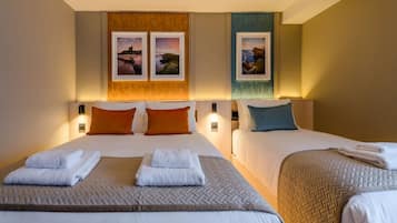 Quarto de hotel com duas camas, cada uma com cabeceira, roupa de cama branca e colcha acolchoada. Há duas molduras de quadros na parede com fotografias de paisagens.