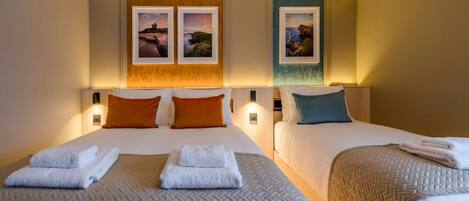 A hotel room with two beds, each with a headboard, white linens, and a quilted bedspread. There are two wall-mounted picture frames with landscape photos.