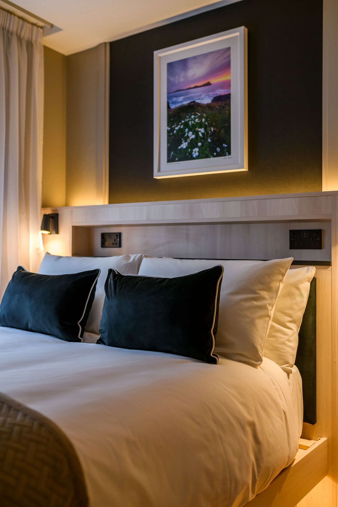Une chambre d’hôtel avec un lit, des oreillers, une table de chevet, un tableau accroché au mur et une fenêtre avec des rideaux.