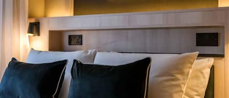 A hotel room with a bed, pillows, a nightstand, a wall-mounted painting, and a window with curtains.