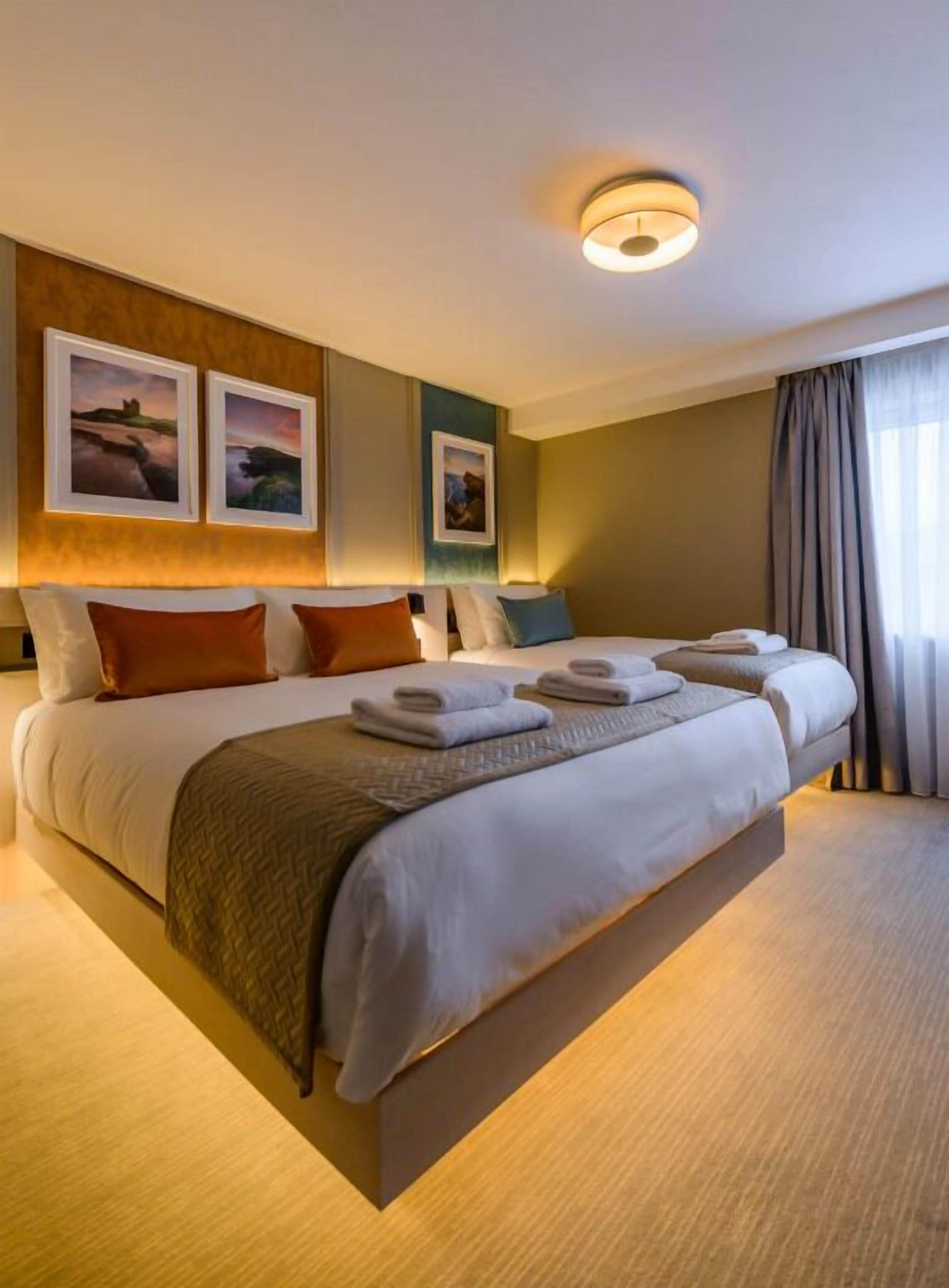 A large bed with multiple pillows and a blanket, a bedside table, and wall-mounted artwork.