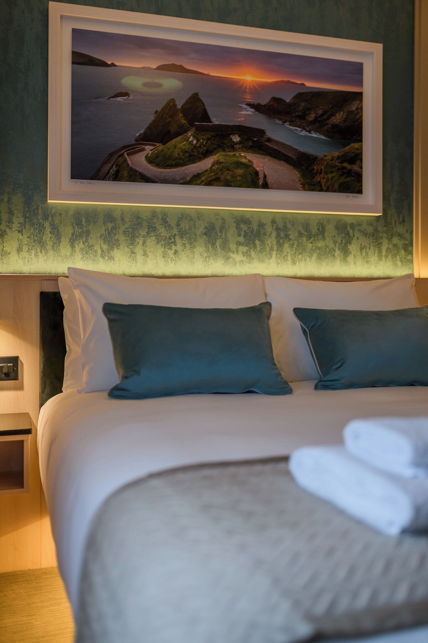 Habitación de hotel con cama, almohadas, un cuadro enmarco de un paisaje y una mesita de noche.